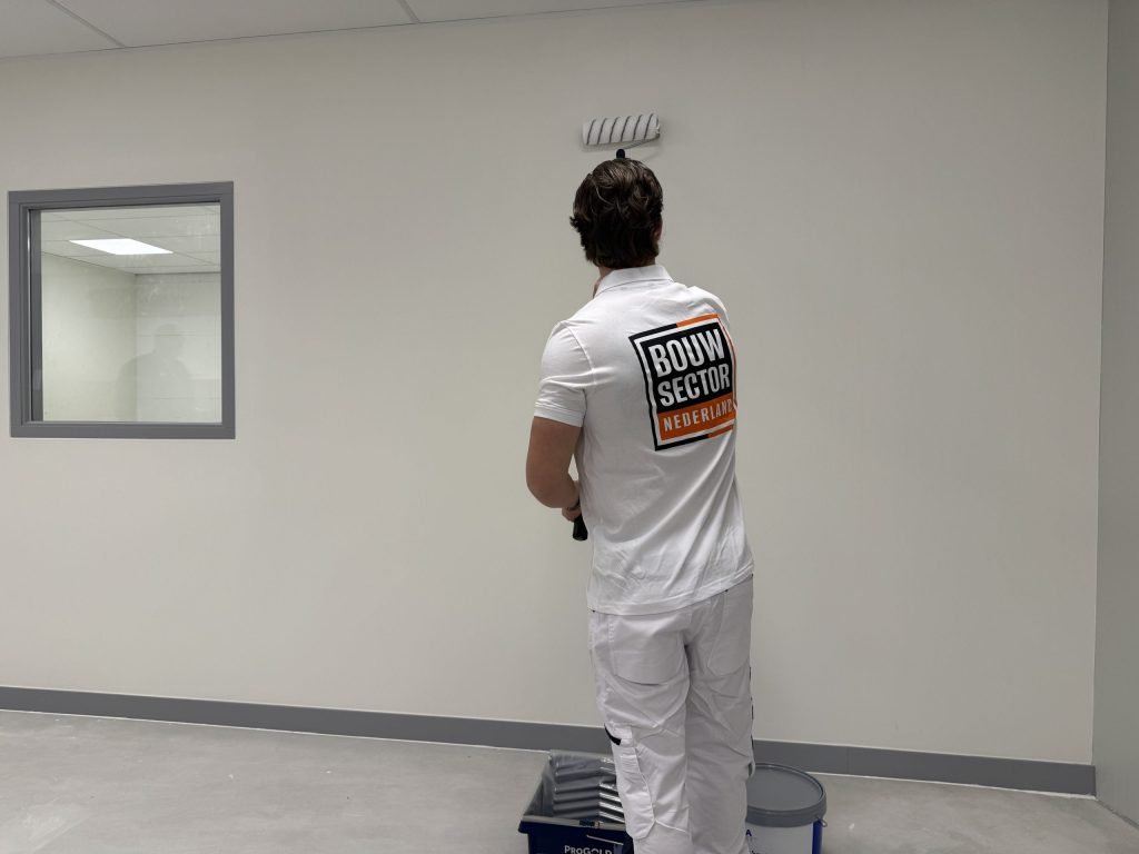 Schilder saust een witte wand boven een verfbak en een emmer muurverf, in een ruimte met een binnenraam