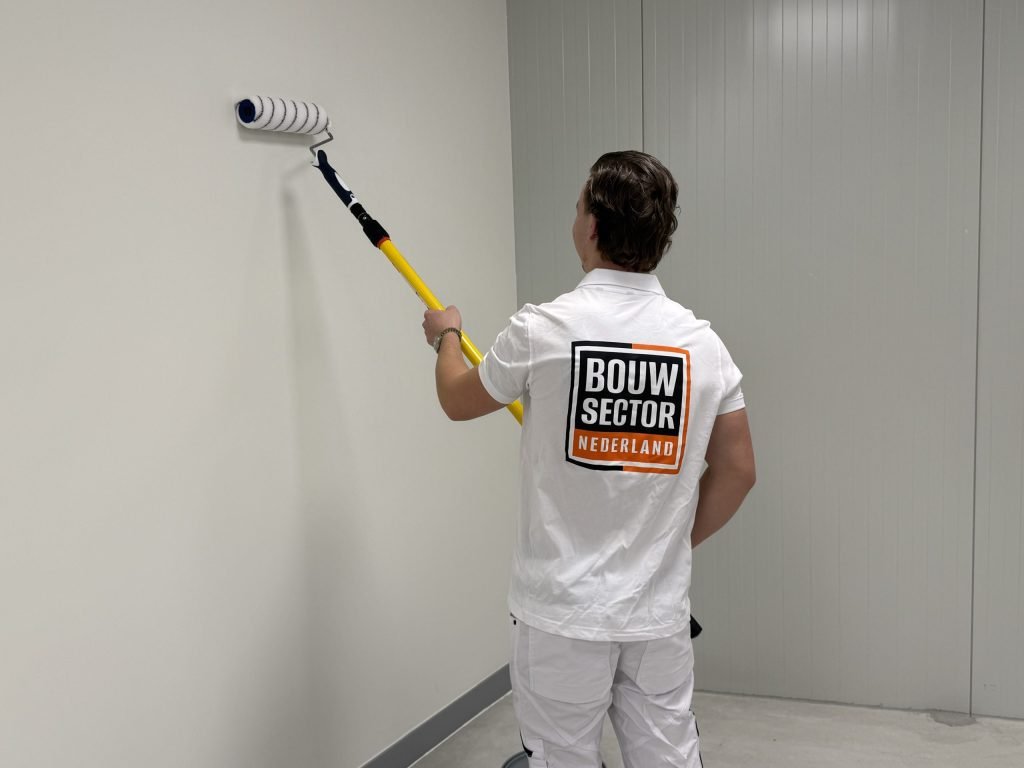 Schilder rolt een lichte wand met een lange roller, naast een wand van metalen panelen