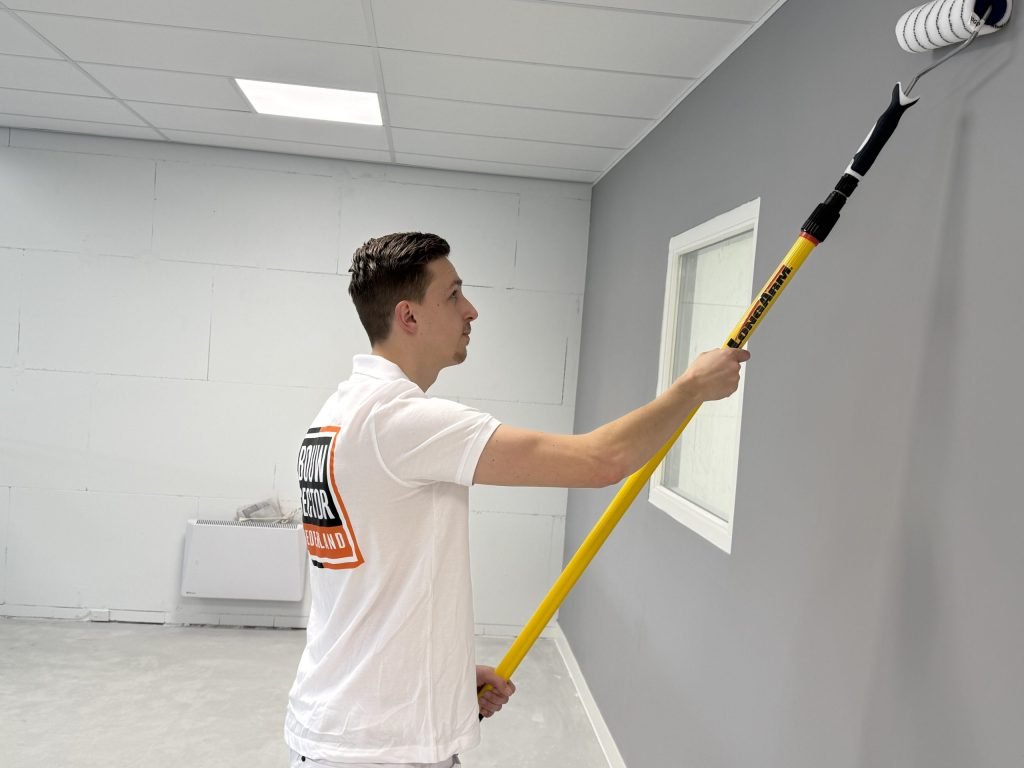 Schilder in wit poloshirt rolt een grijze muur bij een raam, met een kale witte blokkenwand op de achtergrond