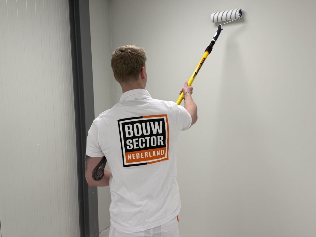 Schilder met een roller aan een gele steel werkt een witte wand bij tot vlak onder het plafond