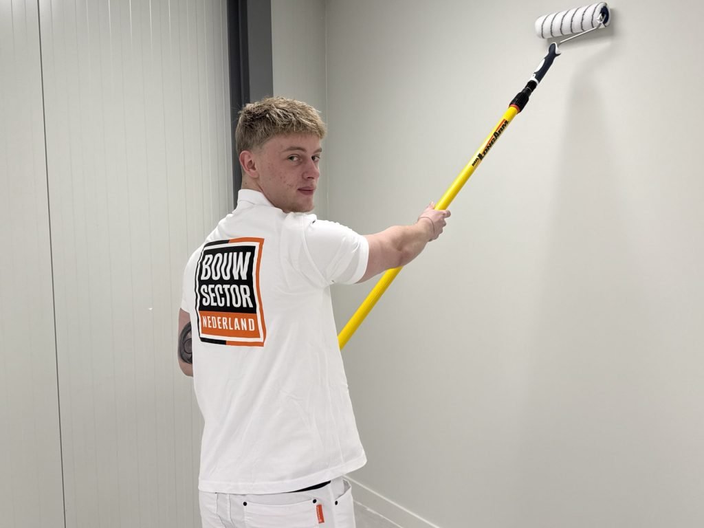 Schilder van Bouwsector Nederland rolt met een verlengsteel een witte muur in een lichte gang