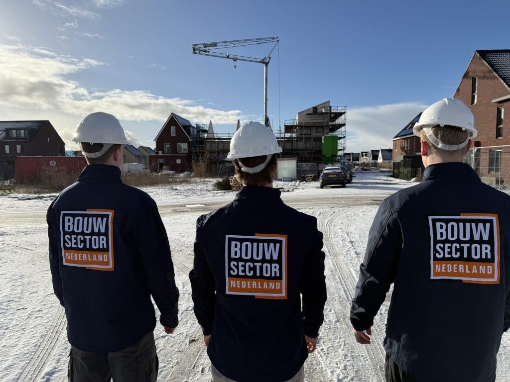 Drie vakmensen van Bouwsector Nederland met witte helm kijken naar een nieuwbouwwijk met een bouwkraan